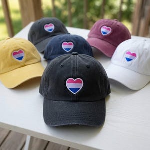 May include: A collection of six baseball caps in yellow, dark gray, navy blue, maroon, white, and black. Each cap features an embroidered heart patch on the front panel, displaying horizontal stripes of pink, purple, and blue colors.
