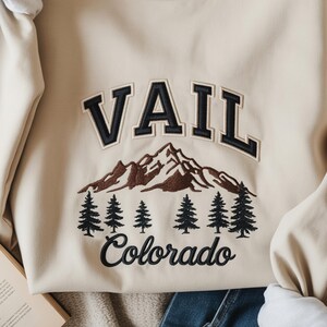 Op de afbeelding: Beige sweatshirt met het woord "VAIL" in marineblauwe gebogen letters boven een berg- en dennenboomafbeelding. Het woord "Colorado" is eronder geborduurd in marineblauw. De sweatshirt heeft lange mouwen.