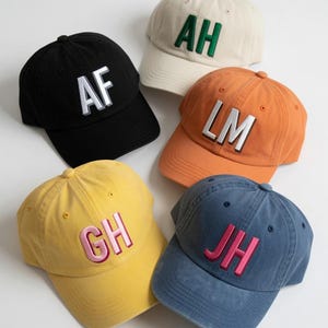 May include: Five baseball caps in black, cream, orange, yellow, and blue. Each cap features two-letter embroidered initials on the front: AF in white, AH in green, LM in white, GH in pink, and JH in pink. The caps have a structured crown and brim.