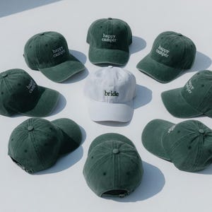 May include: A collection of green baseball caps with the words "happy camper" embroidered in white. One white cap with "bride" embroidered in white is also present. The caps are arranged in a circular pattern.