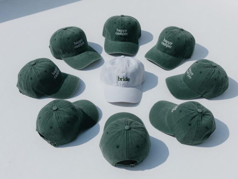 May include: A collection of green baseball caps with the words "happy camper" embroidered in white. One white cap with "bride" embroidered in white is also present. The caps are arranged in a circular pattern.