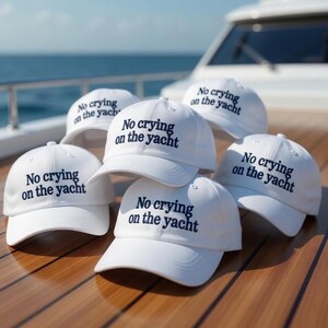 May include: A collection of white baseball caps with the text "No crying on the yacht" in navy blue. The caps are arranged on a wooden surface, with a yacht and the ocean visible in the background.