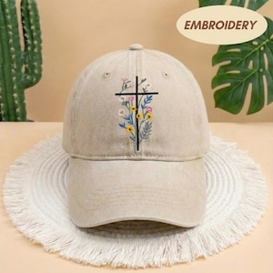 May include: Tan baseball cap with an embroidered black cross design adorned with colorful flowers. The cap features a curved brim and a classic six-panel construction. The word "EMBROIDERY" is printed on a cream-colored oval.