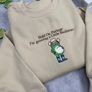 Może przedstawiać: Beżowa bluza z okrągłym dekoltem i brązowym haftowanym napisem "Hold On Partner I'm growing a Little Buckaroo" nad wyhaftowaną zieloną żabą w r&oacute;żowym kowbojskim kapeluszu i niebieskich kowbojkach z białymi gwiazdkami.