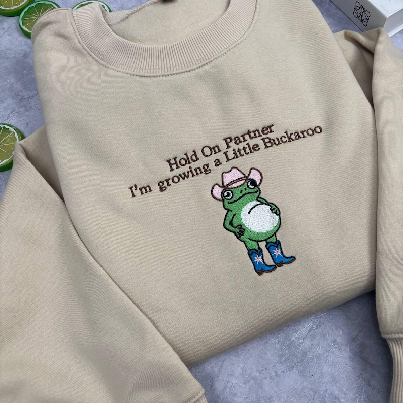 Może przedstawiać: Beżowa bluza z okrągłym dekoltem i brązowym haftowanym napisem "Hold On Partner I'm growing a Little Buckaroo" nad wyhaftowaną zieloną żabą w r&oacute;żowym kowbojskim kapeluszu i niebieskich kowbojkach z białymi gwiazdkami.