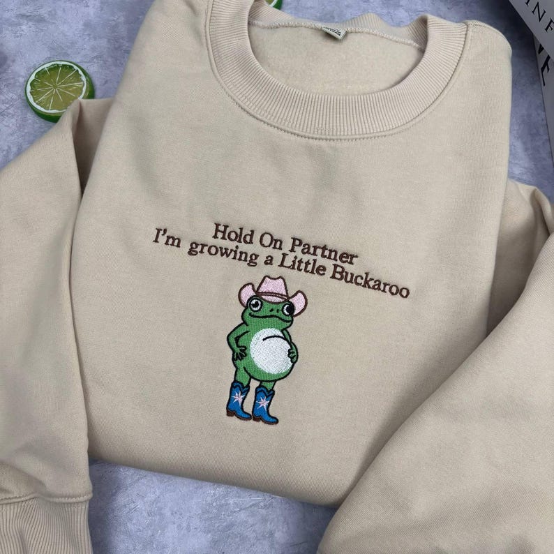Może przedstawiać: Beżowa bluza z okrągłym dekoltem z brązowym haftowanym napisem Hold On Partner I'm growing a Little Buckaroo. Poniżej tekstu znajduje się wyhaftowana zielona żaba w r&oacute;żowym kowbojskim kapeluszu i niebieskich kowbojkach z białymi gwiazdkami.