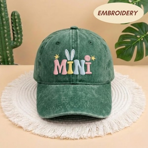 May include: A green baseball cap with the word "MINI" embroidered in colorful letters. The design includes bunny ears and stars. The cap is made of a washed fabric and has a curved brim.