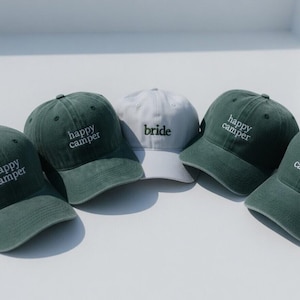 May include: Five baseball caps, four in a muted green and one in white. The green caps have "happy camper" embroidered in white. The white cap has "bride" embroidered in black. All caps have a curved brim.