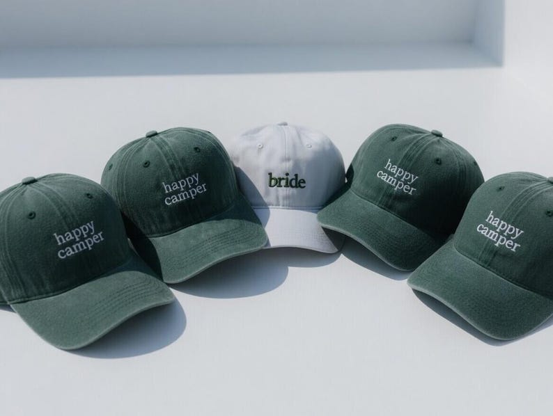 May include: Five baseball caps, four in a muted green and one in white. The green caps have "happy camper" embroidered in white. The white cap has "bride" embroidered in black. All caps have a curved brim.