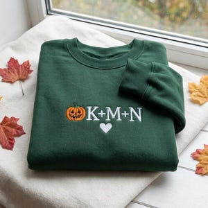 May include: A dark green crewneck sweatshirt featuring an embroidered design on the chest. The embroidery includes an orange jack-o'-lantern, the white text K+M+N, and a small white heart symbol positioned directly below the letters.