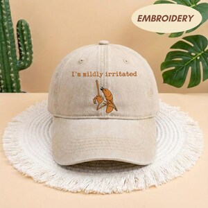 May include: A beige baseball cap with the embroidered phrase "I'm mildly irritated" above a cartoon shrimp holding a stick. The word "EMBROIDERY" is printed on a cream-colored oval on the top right.