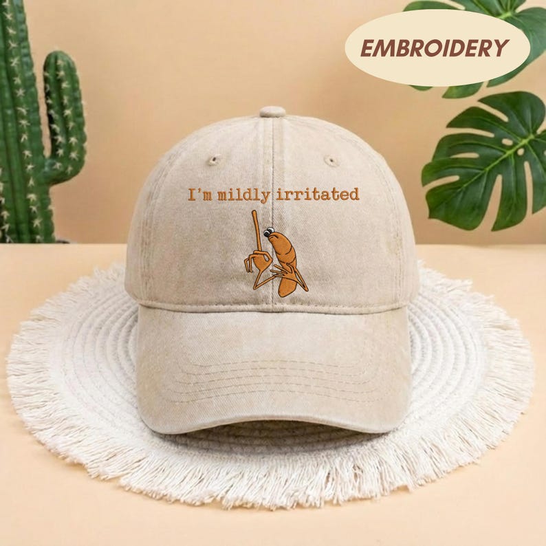 May include: A beige baseball cap with the embroidered phrase "I'm mildly irritated" above a cartoon shrimp holding a stick. The word "EMBROIDERY" is printed on a cream-colored oval on the top right.