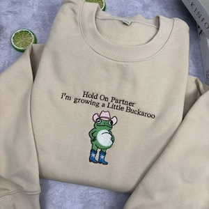 Może przedstawiać: Beżowa bluza z okrągłym dekoltem i ciemnobrązowym haftowanym napisem Hold On Partner I'm growing a Little Buckaroo. Poniżej tekstu znajduje się wyhaftowana zielona żaba w r&oacute;żowym kowbojskim kapeluszu i niebieskich kowbojskich butach.