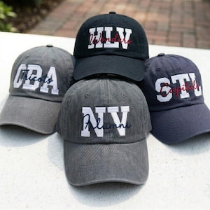 May include: Four baseball caps in black and gray tones, each featuring large white block letters and smaller cursive embroidery. The caps display the text HLV Wonders, OBA Tricots, NV Alumni, and STL Capitals, with each hat having a curved brim.