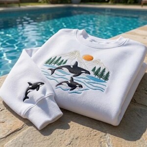May include: A white crewneck sweatshirt featuring an embroidered design of two orcas swimming in blue water, with green pine trees, a mountain range, and an orange sun in the background. A single orca is embroidered on the left sleeve cuff.
