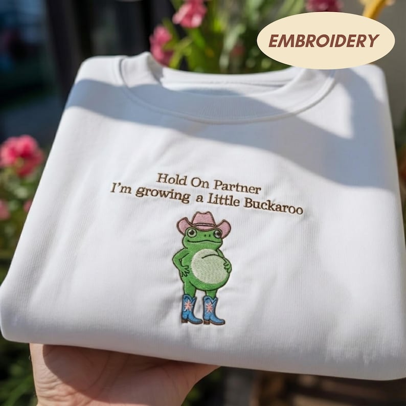 Może przedstawiać: Biała bluza z okrągłym dekoltem z brązowym haftowanym napisem "Hold On Partner I'm growing a Little Buckaroo" nad zieloną żabą w r&oacute;żowym kowbojskim kapeluszu i niebieskich kowbojkach z białym detalem w kształcie gwiazdy z przodu.