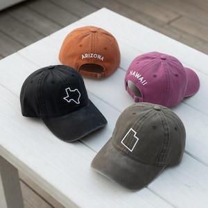 May include: Four baseball caps in various colors: black, orange, purple, and olive green. Each cap features a state outline or state name in white. The caps are arranged on a white wooden surface.