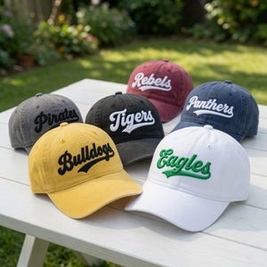May include: A collection of six baseball caps in various colors including yellow, white, grey, black, maroon, and navy blue. Each cap features embroidered team names in script font, specifically Bulldogs, Eagles, Pirates, Tigers, Rebels, and Panthers.
