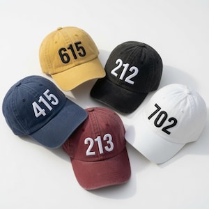 May include: Five baseball caps in yellow, black, blue, maroon, and white. Each cap features a distinct three-digit number embroidered on the front: 615 in black, 212 in white, 415 in white, 213 in white, and 702 in black. All caps have a curved brim.