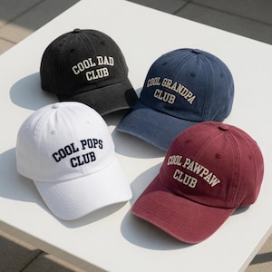 May include: Four baseball caps in black, navy, white, and burgundy. Each cap has the words "COOL DAD CLUB", "COOL GRANDPA CLUB", "COOL POPS CLUB", and "COOL PAWPAW CLUB" embroidered in white. The caps are arranged on a white surface.