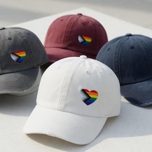 May include: A collection of baseball caps in various colors: gray, burgundy, navy, and white. Each cap features an embroidered heart with a rainbow LGBTQ+ pride flag design. The caps are made of a soft, worn material.
