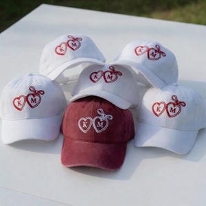 May include: A collection of baseball caps, primarily white, with a heart-shaped design featuring the letters "K" and "M" in red. One cap is a deep red color. The caps are arranged on a white surface.