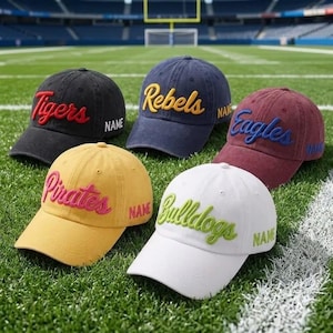 May include: Five baseball caps in black, navy blue, maroon, yellow, and white. Each cap features embroidered team names in contrasting colors: Tigers, Rebels, Eagles, Pirates, and Bulldogs. The word NAME is embroidered on the side of each cap.