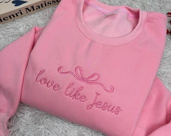 May include: A light pink sweatshirt with the embroidered phrase "love like Jesus" in a cursive font. The sweatshirt has a crew neck and long sleeves. The embroidery includes a bow design above the words.