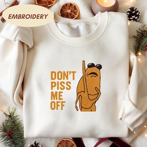 May include: Cream-colored sweatshirt with the embroidered text "DON'T PISS ME OFF" and a cartoon character wearing sunglasses. The character is a light brown color. The word "EMBROIDERY" is printed on the top left of the sweatshirt.