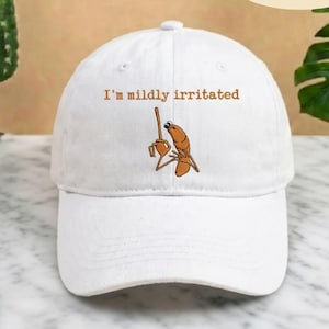 May include: White baseball cap with the text "I'm mildly irritated" in brown lettering. The cap features an embroidered cartoon shrimp character with a raised finger. The cap is made of a soft fabric and has a curved brim.