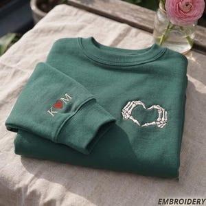 May include: A dark green crewneck sweatshirt featuring white embroidered skeleton hands forming a heart shape on the chest. The sleeve cuff displays the embroidered letters K and M separated by a small red heart. The fabric has a ribbed neckline.