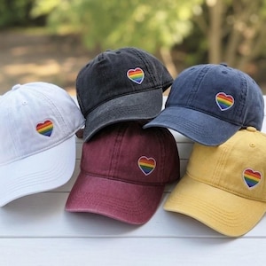 May include: A collection of baseball caps in various colors: white, black, navy, burgundy, and yellow. Each cap features a rainbow heart patch on the front. The caps are arranged on a white wooden surface.