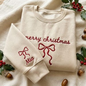 May include: A cream-colored crewneck sweatshirt featuring red embroidered text that reads merry christmas, accompanied by a red embroidered bow design on the chest and a matching red bow with the name Mia embroidered on the sleeve cuff.