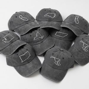 May include: A collection of gray baseball caps, each embroidered with a white outline of a U.S. state. The caps are arranged in a pile, showcasing various state designs. The caps have a vintage, worn look.