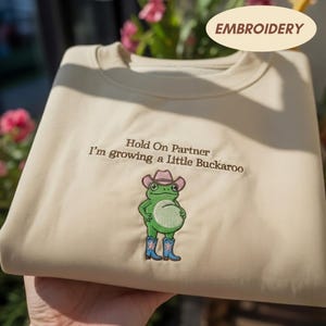 Może przedstawiać: Beżowa bluza z okrągłym dekoltem i brązowym haftem z napisem "Hold On Partner I'm growing a Little Buckaroo" nad zieloną żabą w r&oacute;żowym kowbojskim kapeluszu i niebieskich kowbojkach z białym wzorem gwiazdy z przodu.
