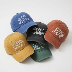 May include: A collection of five baseball caps in blue, mustard yellow, charcoal gray, dark green, and terracotta. Each cap features white embroidered airport code lettering: ATL, DEN, ORD, SFO, and AUS, with a structured crown and curved brim.