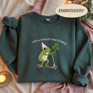 May include: Dark green sweatshirt with the embroidered phrase "Off to commit Irish foolery." The design features a green frog riding a scooter and holding a bouquet of shamrocks. The word "EMBROIDERY" is printed on a cream-colored oval.