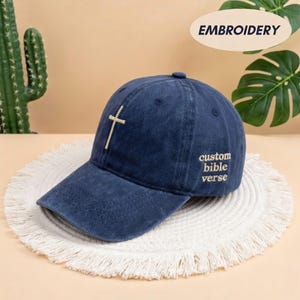 May include: A navy blue baseball cap with a curved brim. The cap features a stitched cross on the front and the words "custom bible verse" embroidered on the side. The cap is sitting on a white, fringed mat.
