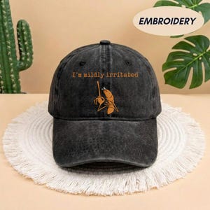 May include: A black baseball cap with the embroidered phrase "I'm mildly irritated" above a cartoonish image of a shrimp holding a stick. The cap is sitting on a white, fringed placemat.