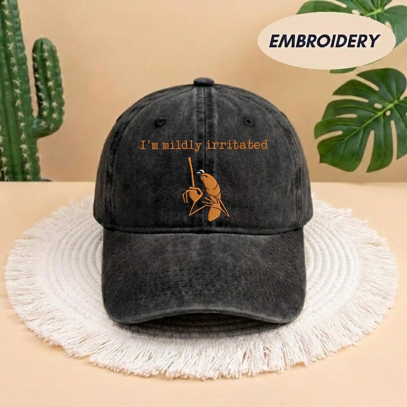 May include: A black baseball cap with the embroidered phrase "I'm mildly irritated" above a cartoonish image of a shrimp holding a stick. The cap is sitting on a white, fringed placemat.