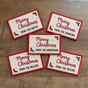 May include: Five rectangular off-white fabric gift tags with red embroidered Merry Christmas text and green embroidered holly accents. Each tag features a unique family name printed in black text following the word FROM.
