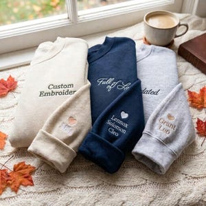 May include: Three crewneck sweatshirts in cream, navy blue, and heather gray. Each features custom embroidery on the chest and sleeve, including names like Mia, Lennox, Solomon, Cleo, Grant, and Leo, accompanied by small embroidered heart icons.