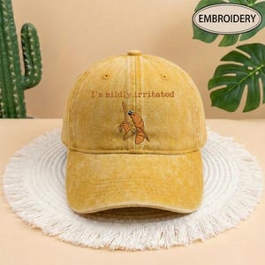 May include: Mustard yellow baseball cap with the embroidered phrase "I'm mildly irritated" above a cartoon shrimp holding a tiny broom. The cap has a curved brim and a distressed look, perfect for casual wear.