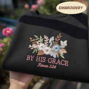 May include: Black sweatshirt with floral embroidery in white, blue, and tan. The text "BY HIS GRACE Romans 3:24" is embroidered in pink. The word "EMBROIDERY" is in a tan oval.