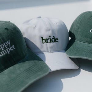 May include: Three baseball caps. Two are dark green with the embroidered text "happy camper". The third is white with the embroidered word "bride" in green. All caps have a curved brim and adjustable strap.