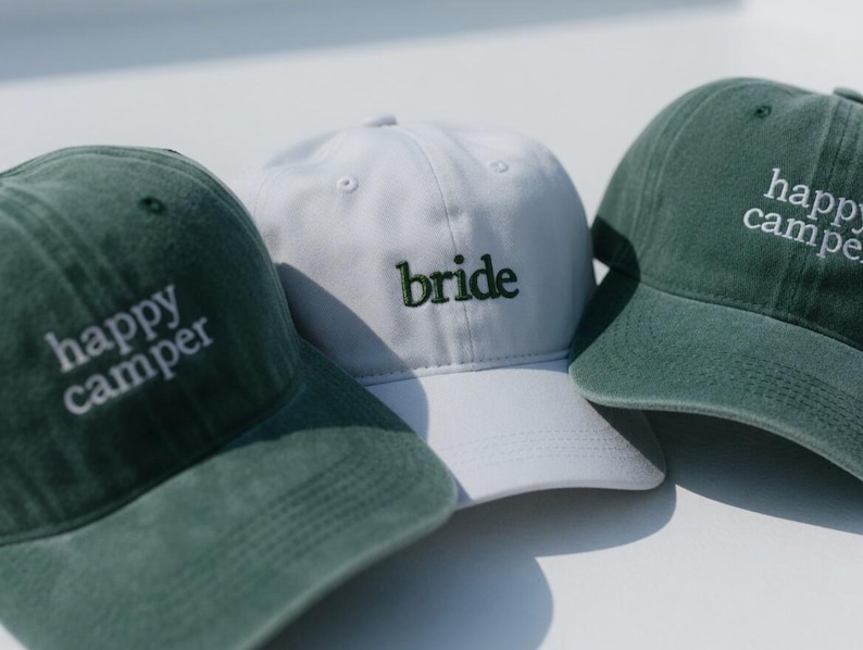 May include: Three baseball caps. Two are dark green with the embroidered text "happy camper". The third is white with the embroidered word "bride" in green. All caps have a curved brim and adjustable strap.