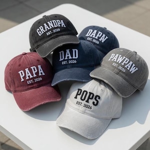May include: A collection of six baseball caps in various colors, each embroidered with a different family-related title in white, such as "GRANDPA," "DAD," and "POPS." The text "EST. 2026" is below each title. The caps have a distressed, vintage look.