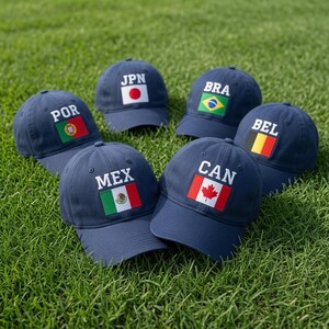 May include: Six navy blue baseball caps arranged in a circle, each featuring a white embroidered country code and a corresponding national flag patch. The flags represent Portugal, Japan, Brazil, Belgium, Mexico, and Canada, with white text labels.