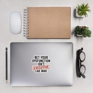 May include: A laptop sticker with the text "BET YOUR DYSFUNCTION ISN'T EXECUTIVE LIKE MINE" in black and red. The sticker is on a laptop, with a mouse, pen, notebook, glasses, and small plants nearby.