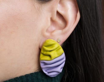 May include: A close-up of a two-tone earring. The top half is yellow, and the bottom half is lavender. The earring has a textured, rippled appearance. The earring is worn on the ear of a person wearing a dark green sweater.